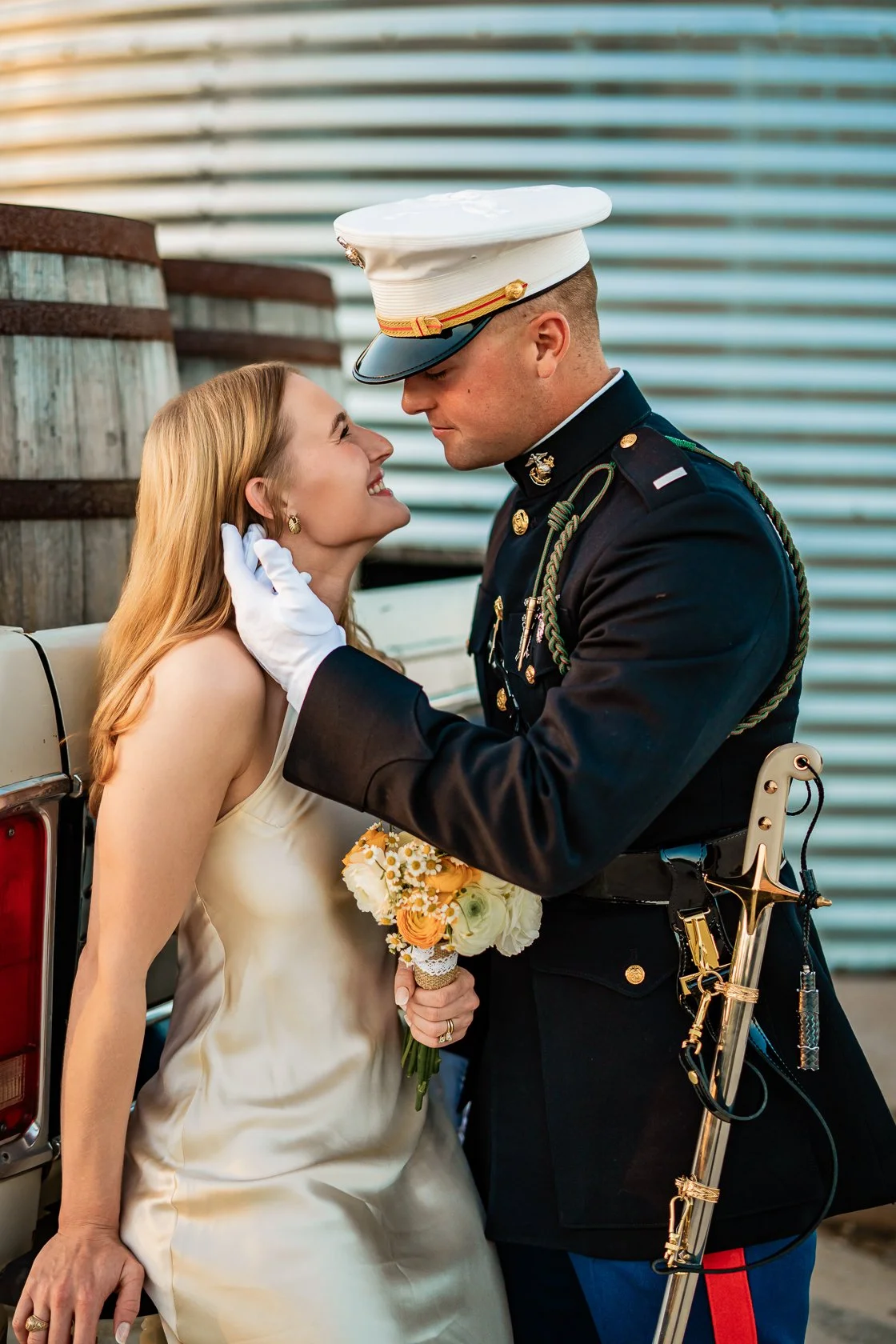 A bride and a soldier in uniform share a tender moment, close together with the soldier holding the bride's face, while she smiles and holds a bouquet of flowers, outdoors near a vehicle and large industrial building.