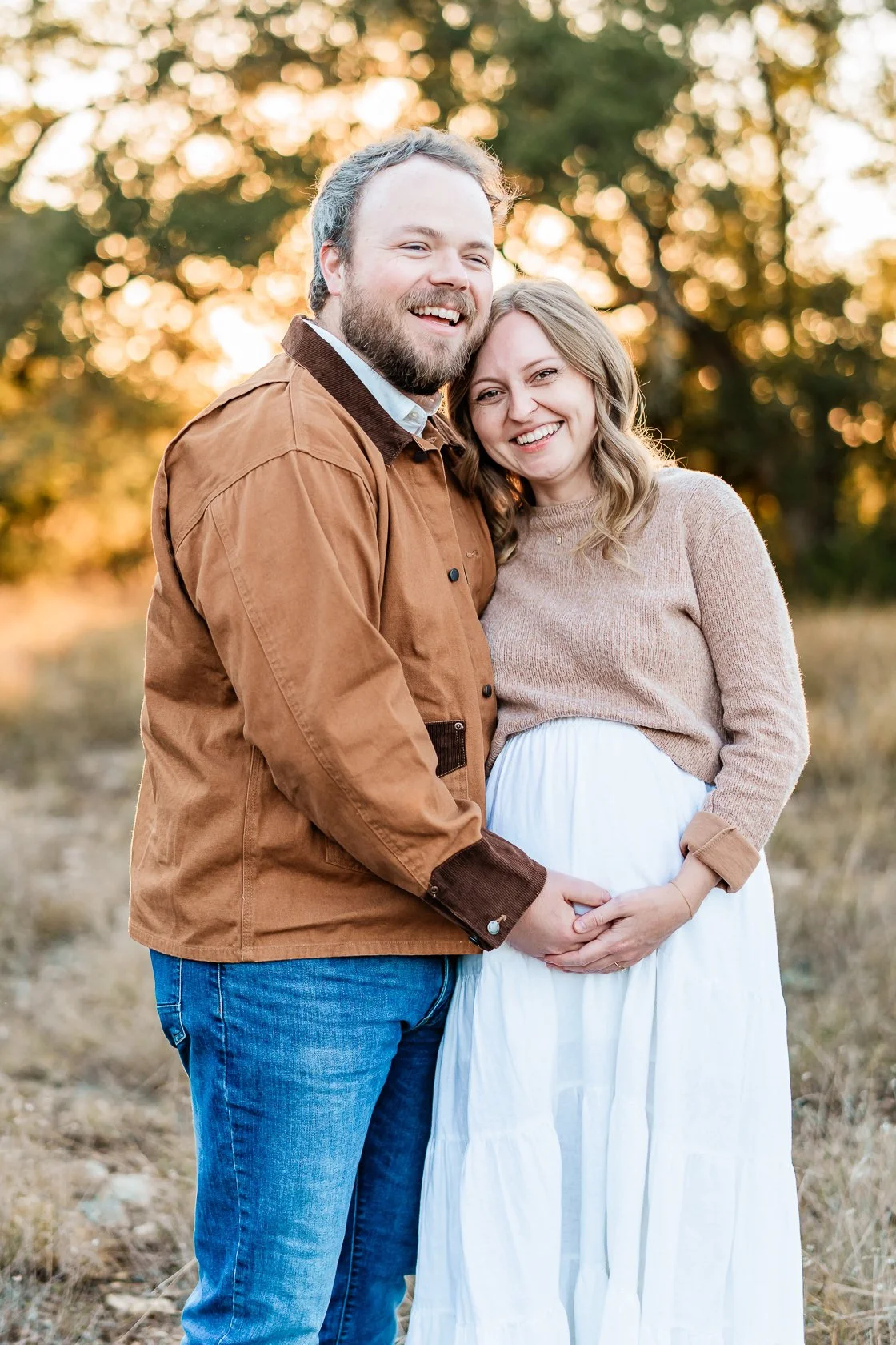 A happy couple, with the woman pregnant, standing outdoors during sunset, holding hands and smiling.