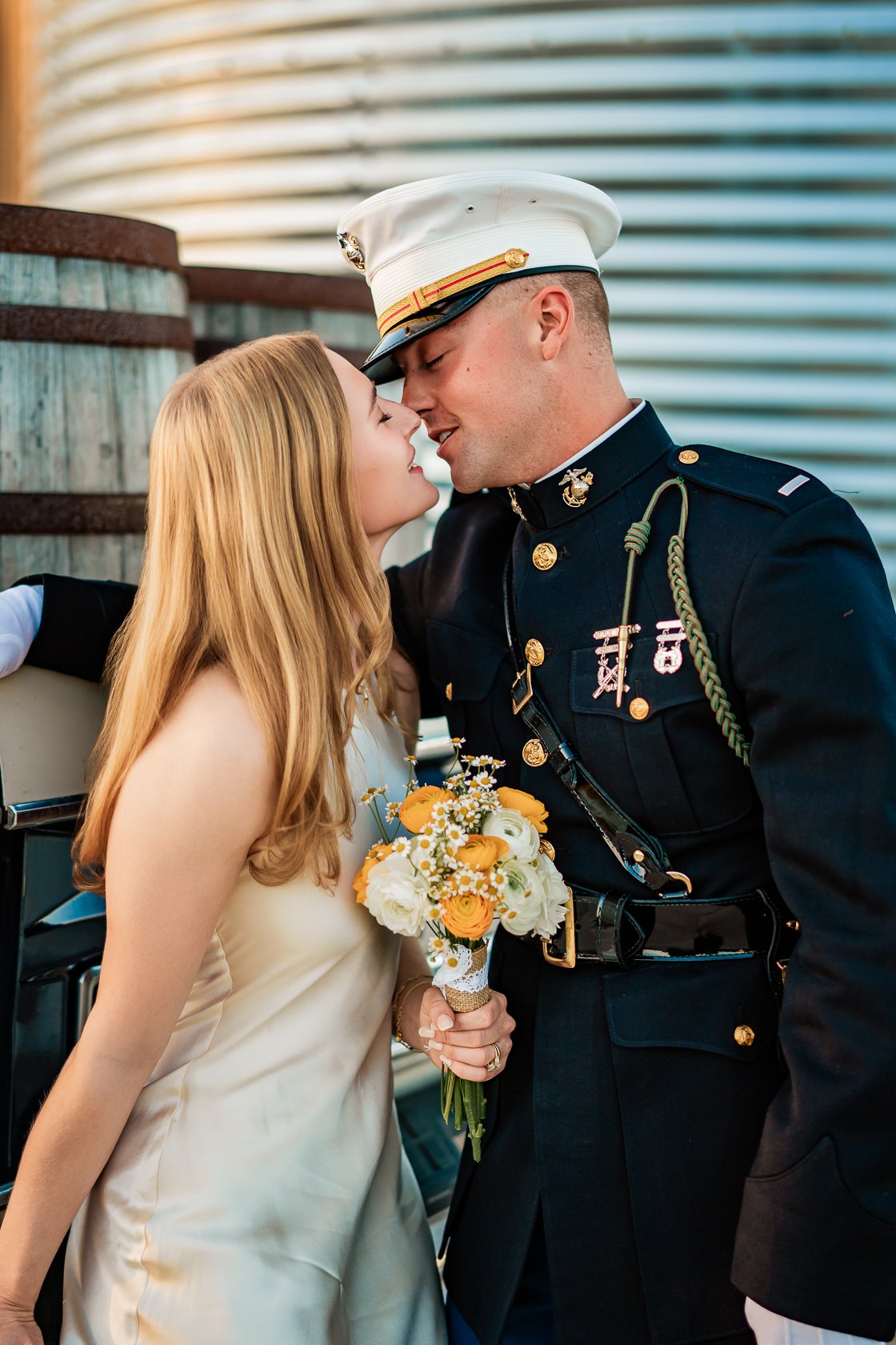 A woman in a white dress holding a bouquet of yellow and white flowers stands close to a man in a US Marine dress uniform, both leaning in with their foreheads touching, sharing an intimate moment.