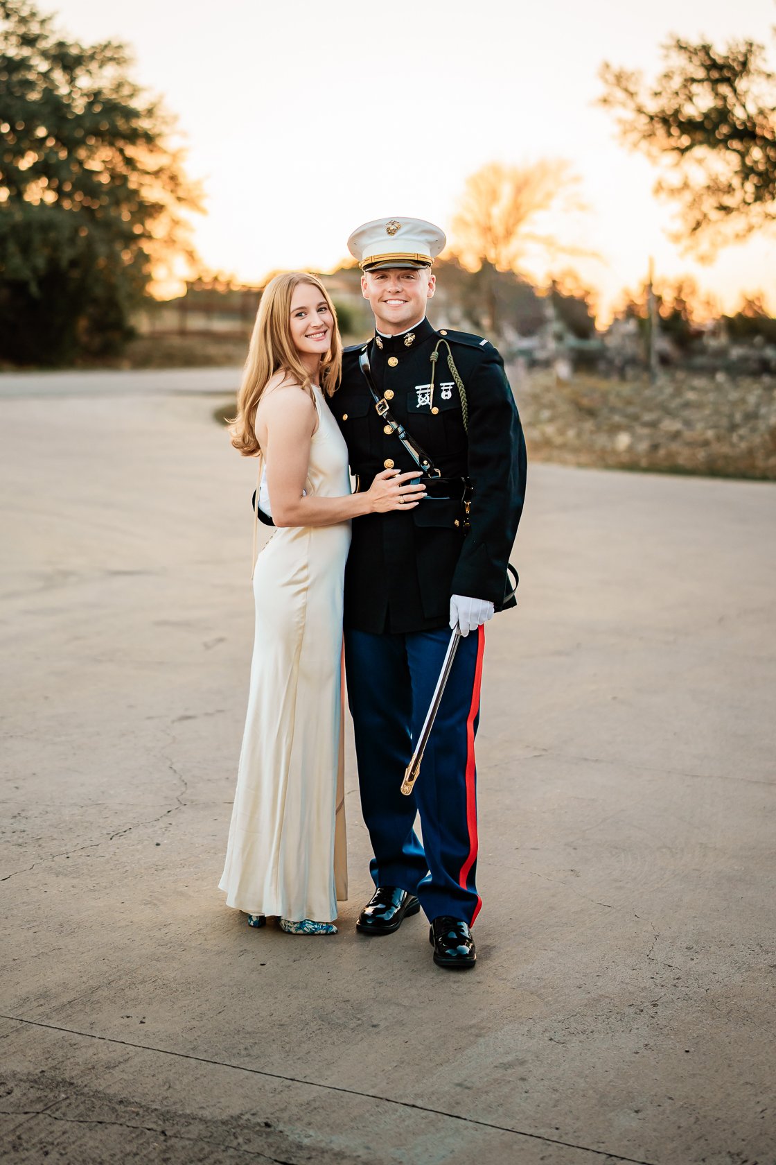 A woman in a white dress and a man in a military uniform standing together outdoors at sunset