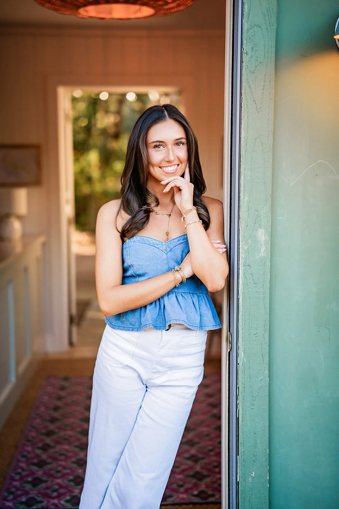 A young woman with long dark hair, wearing a blue sleeveless top and white pants, standing next to a glass door inside a house, smiling at the camera.