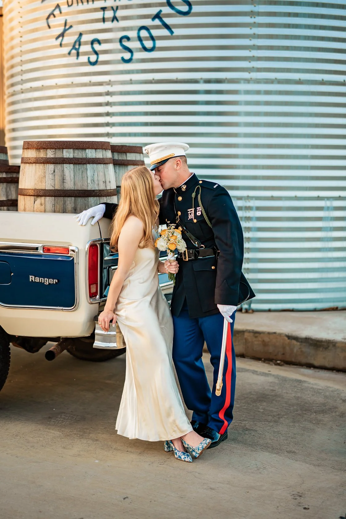 A couple, dressed in wedding attire, sharing a kiss in front of a vintage pickup truck with barrels in the back. The groom is in a U.S. military uniform, and the bride is holding a bouquet of flowers.