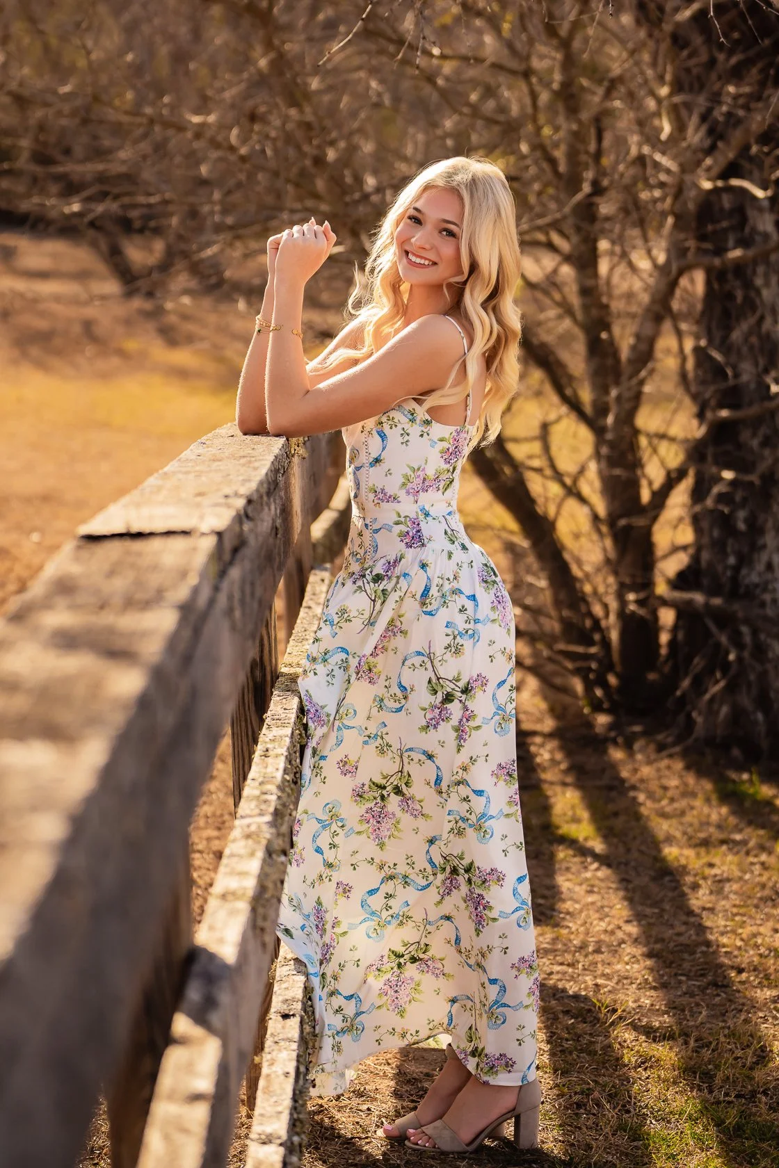 A young woman with blonde hair in a floral dress standing outdoors by a wooden fence, smiling and looking at the camera during golden hour.