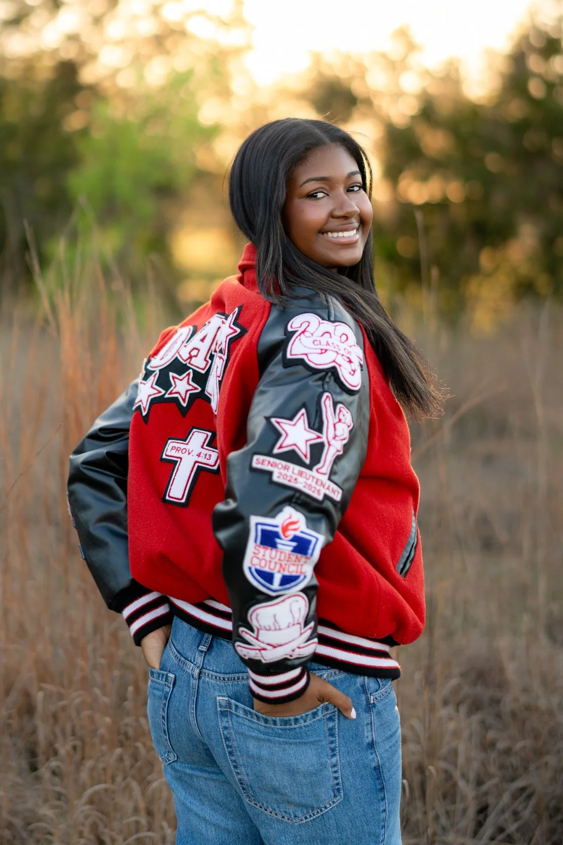 A smiling young woman with long black hair wearing a red varsity jacket with patches and blue jeans standing outdoors in a field with tall grass during sunset.