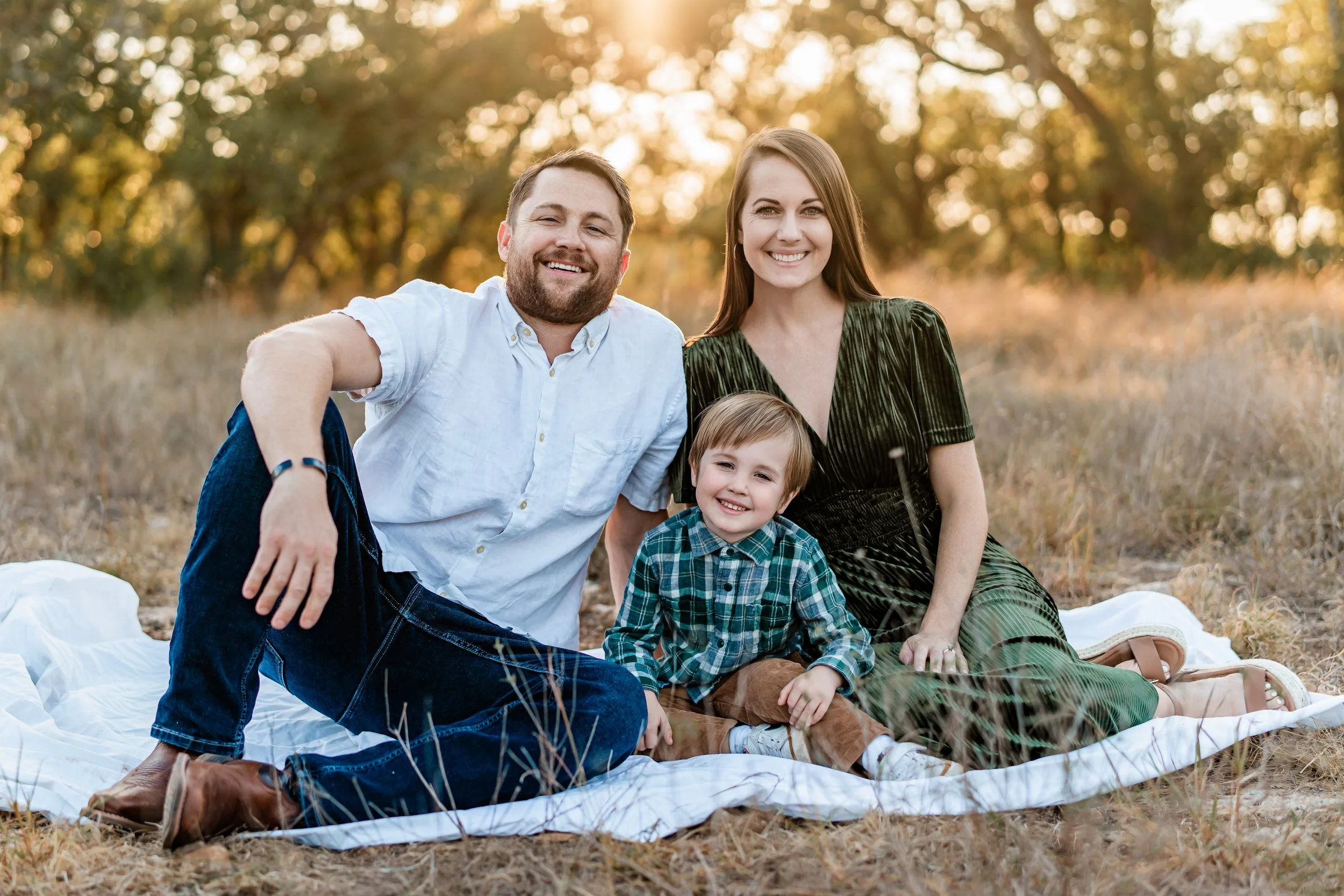 A happy family of three sitting on a white blanket outdoors during sunset, with trees in the background.