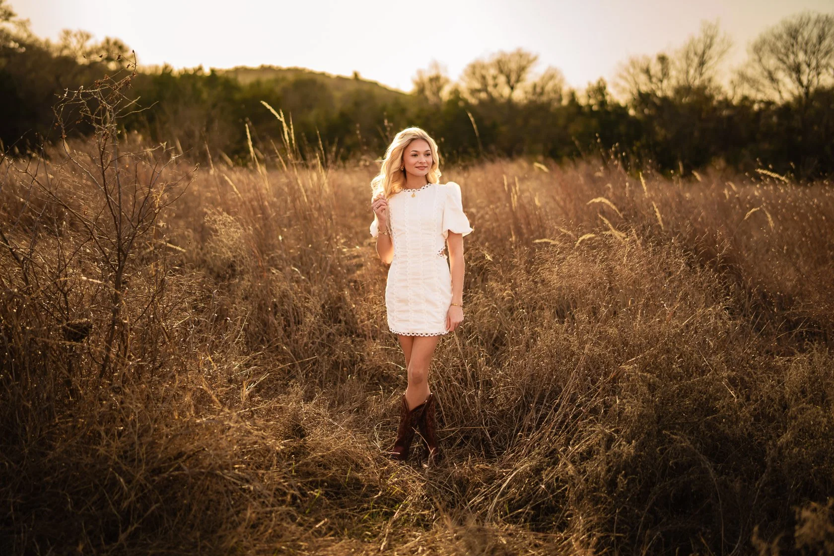 A woman in a white dress and cowboy boots standing in a field at sunset.