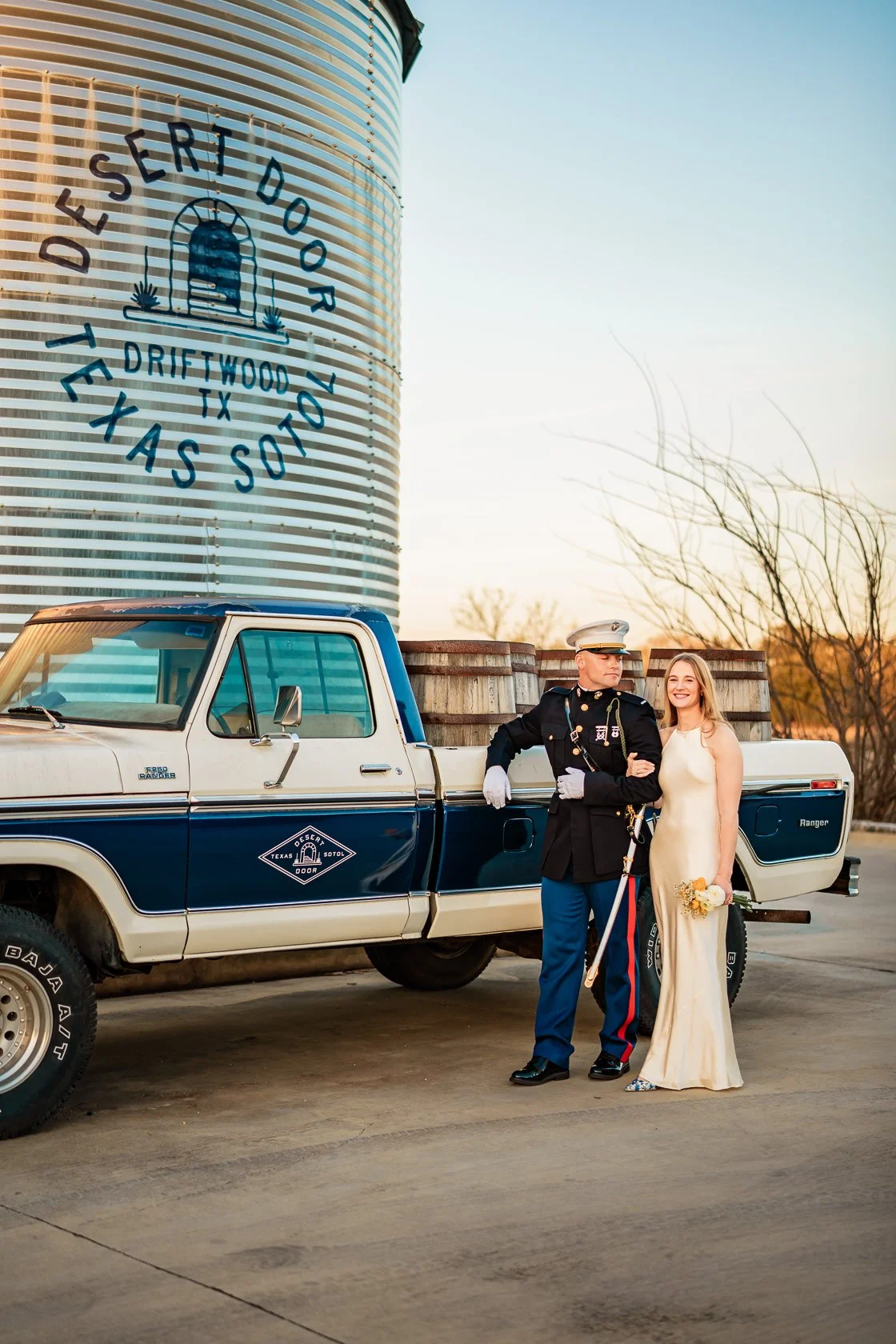 A bride and a military soldier standing beside a vintage Texas Ranger pickup truck with a water tower in the background. The water tower has the text 'Desert Door, Driftwood TX, Texas Sol't' and an illustration of a door with a handle and sun rays.