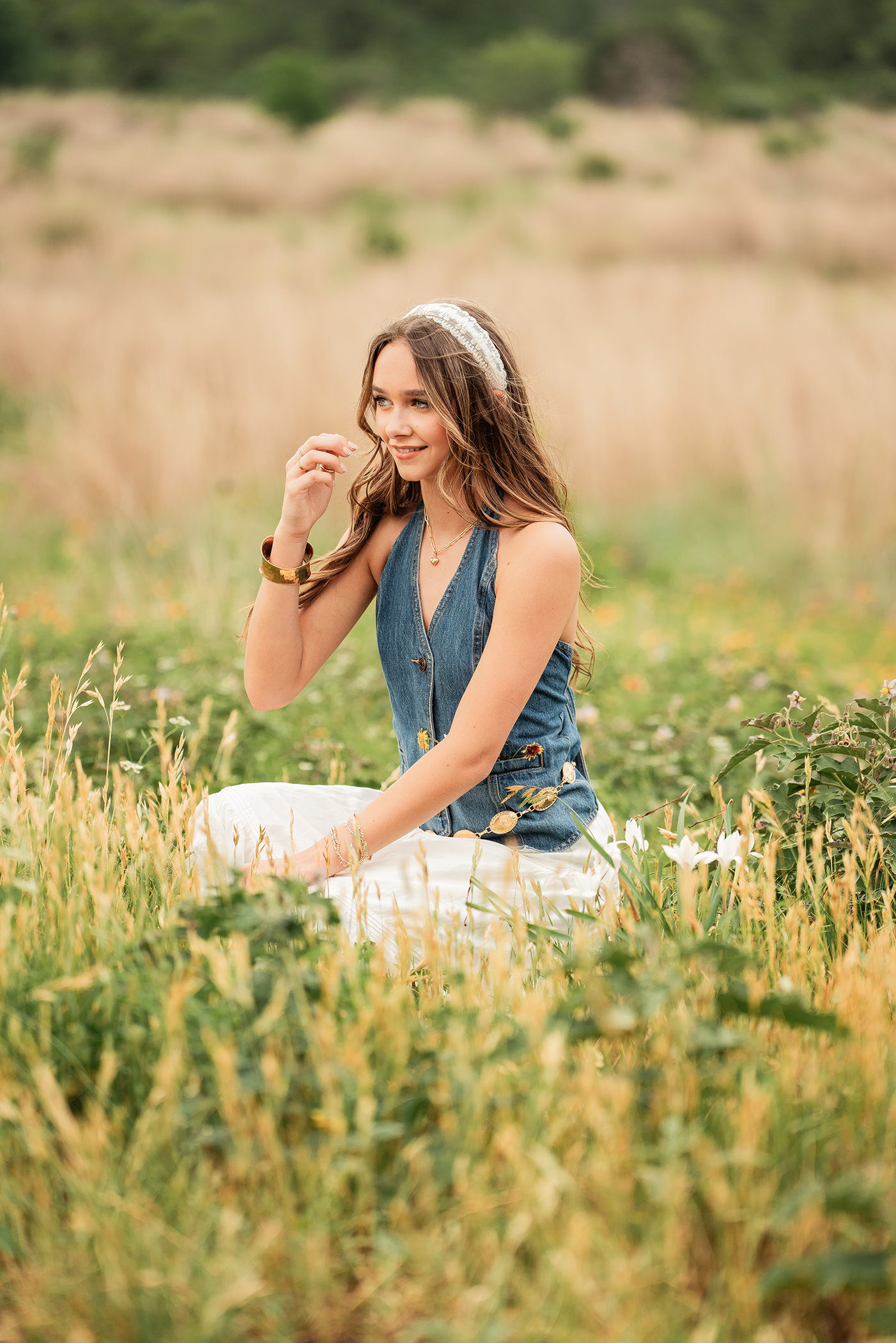 A young woman with long wavy brown hair sitting in a grassy field with wildflowers, wearing a sleeveless denim vest, white pants, a lace headband, and jewelry, smiling and looking to the side.