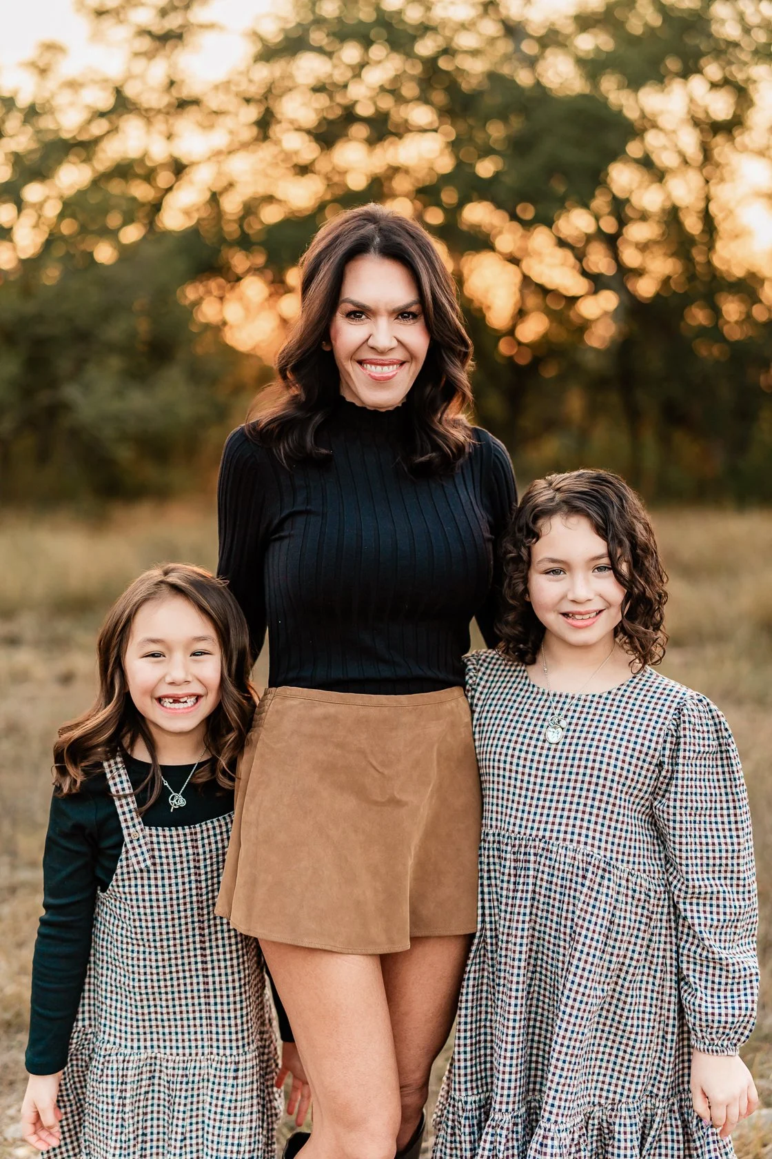 A woman and two young girls standing outdoors during sunset, smiling at the camera.