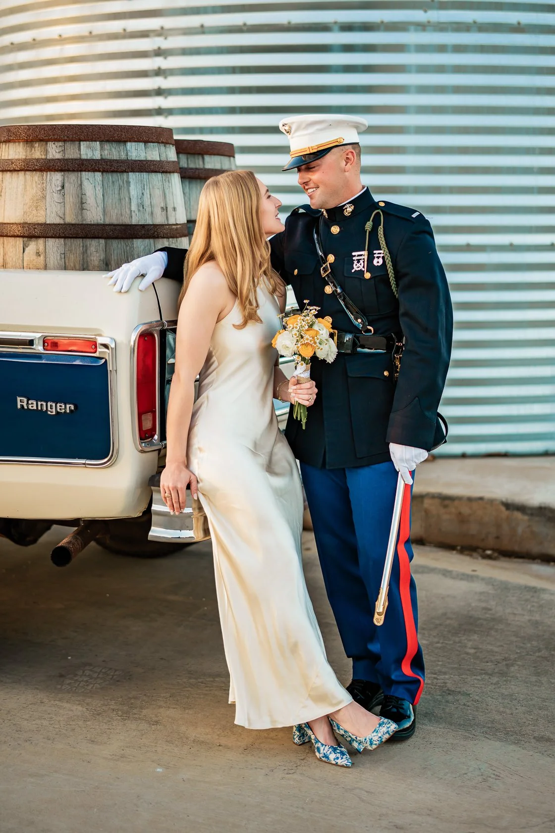 A couple dressed in wedding attire standing beside a military officer in uniform, holding a bouquet of flowers, in front of a vintage truck with a modern building in the background.