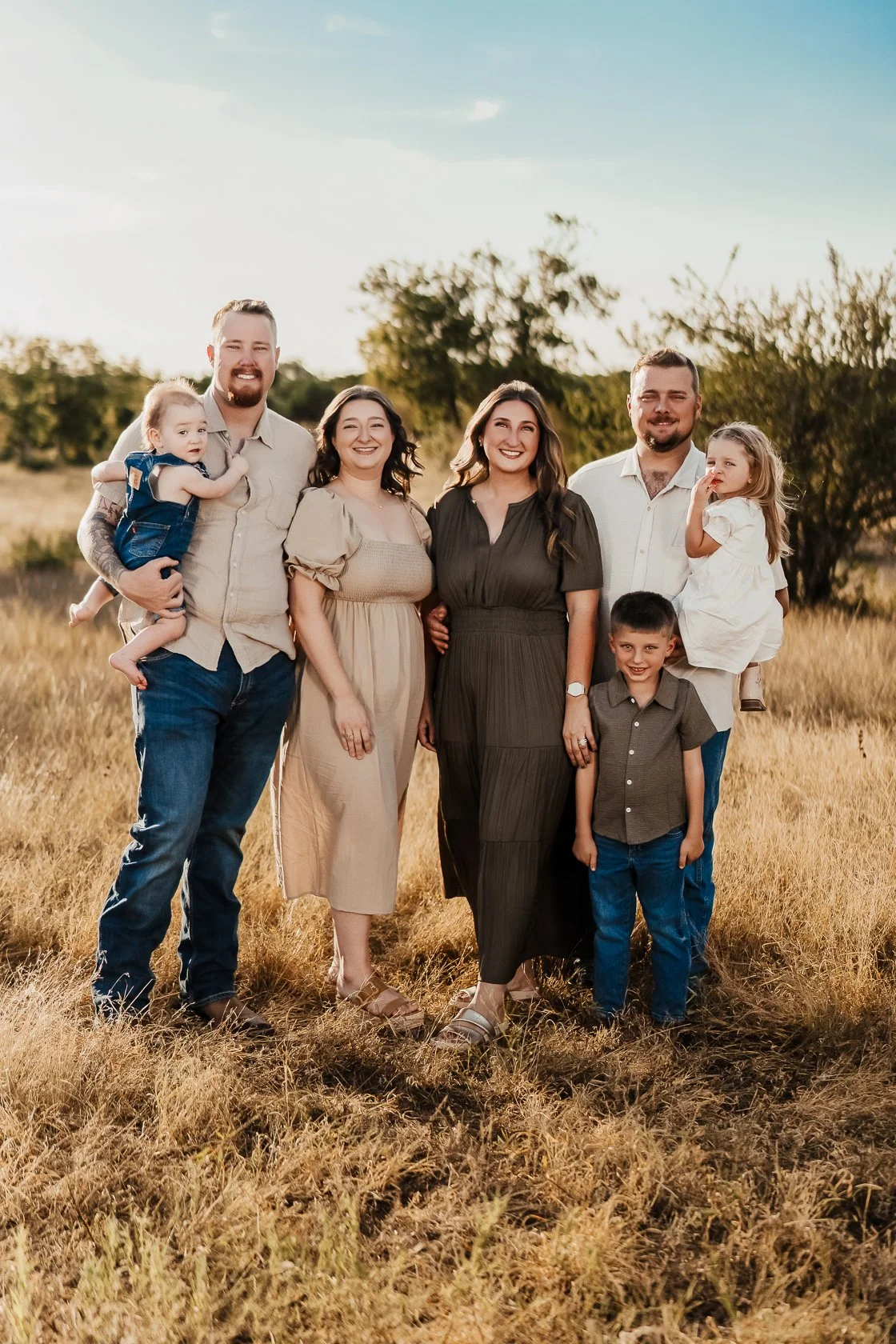 A family of seven standing together outdoors in a field, smiling at the camera during golden hour, with trees and a clear sky in the background.