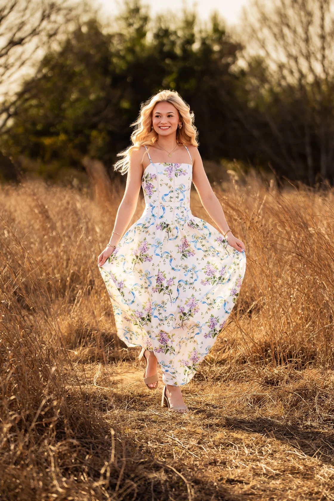 A woman with blonde hair wearing a floral maxi dress, walking through a field of tall, dry grass during sunset.