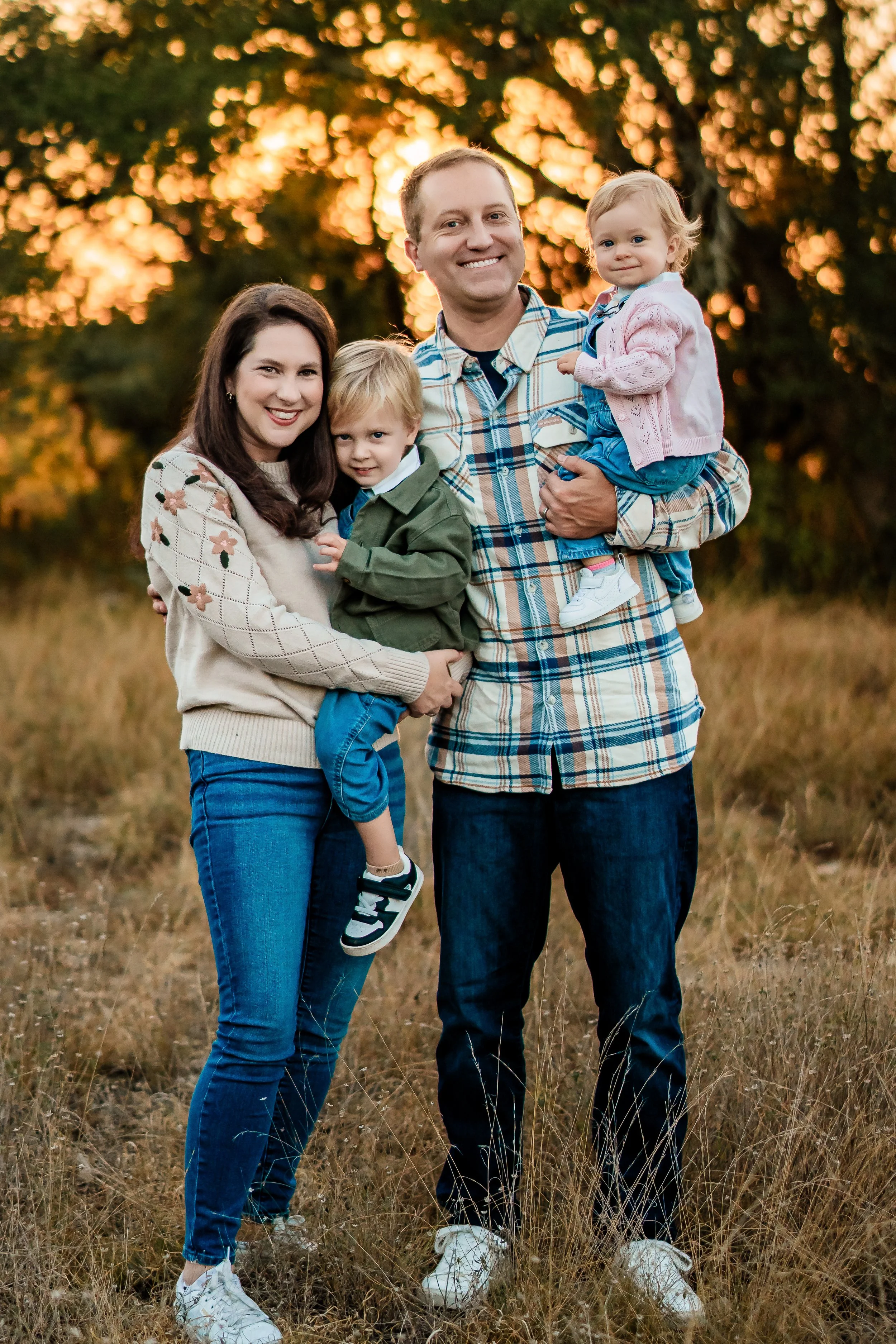 A family of four standing outdoors in a grassy field at sunset, smiling at the camera. The mother has dark brown hair, wearing a cream sweater and blue jeans. The father has blond hair, wearing a plaid shirt and dark jeans, holding a young girl with 