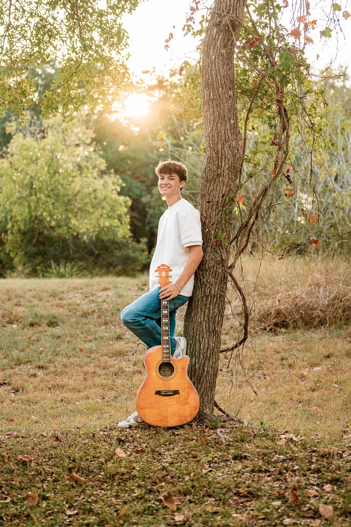 A young man leaning against a tree outdoors during sunset, holding an acoustic guitar, wearing a white shirt and blue jeans, smiling at the camera.