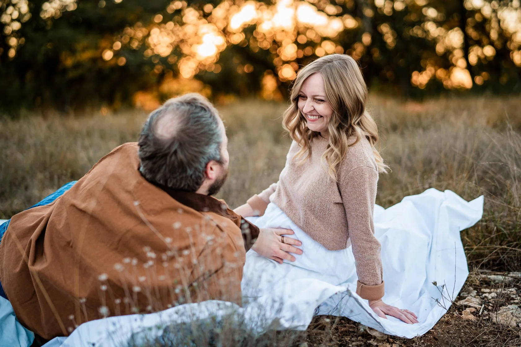 A man kneeling beside a pregnant woman on the ground outdoors during sunset, smiling at each other with trees and orange sky in the background.