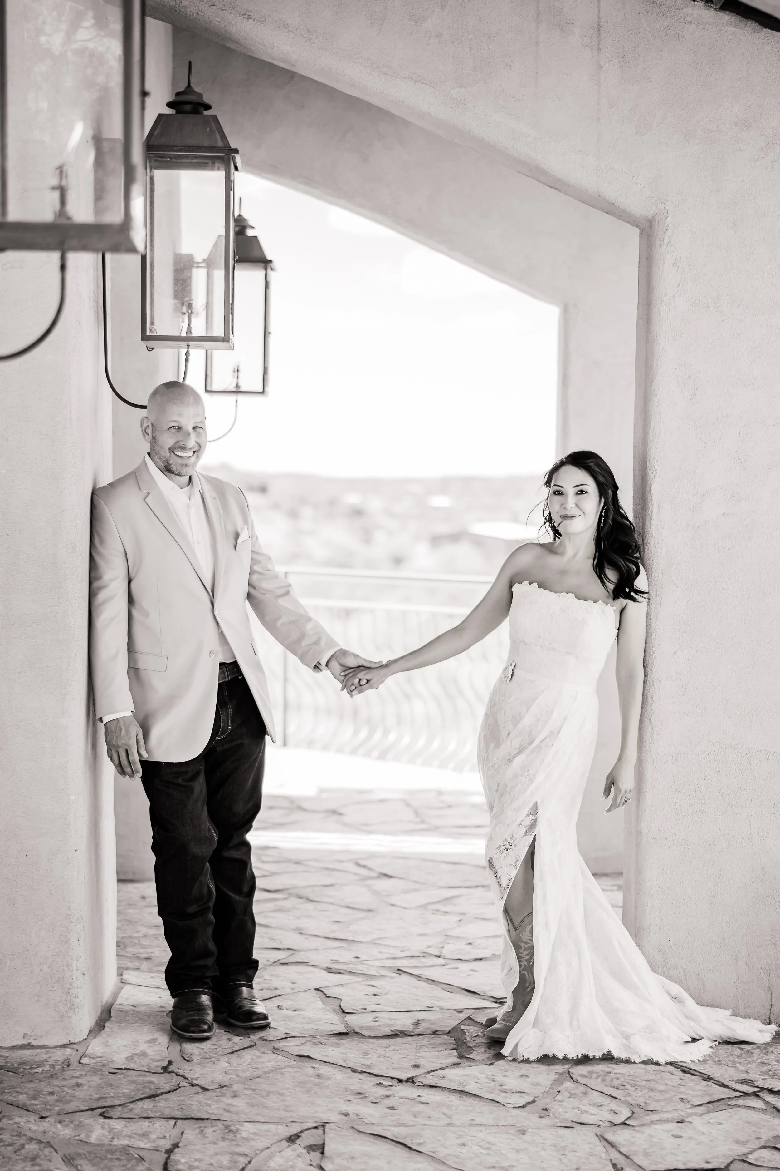 A black-and-white photo of a bride and a man, possibly her father, holding hands and smiling. The bride is wearing a strapless wedding gown, and the man is in a light-colored suit jacket and dark pants. They are standing under a covered outdoor space