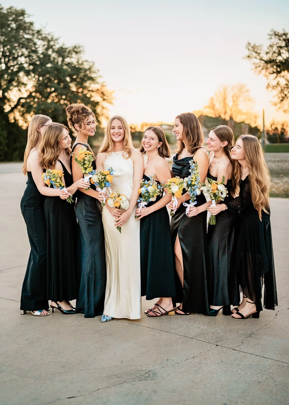 Group of women in black dresses and one woman in a white dress holding bouquets outside during sunset.