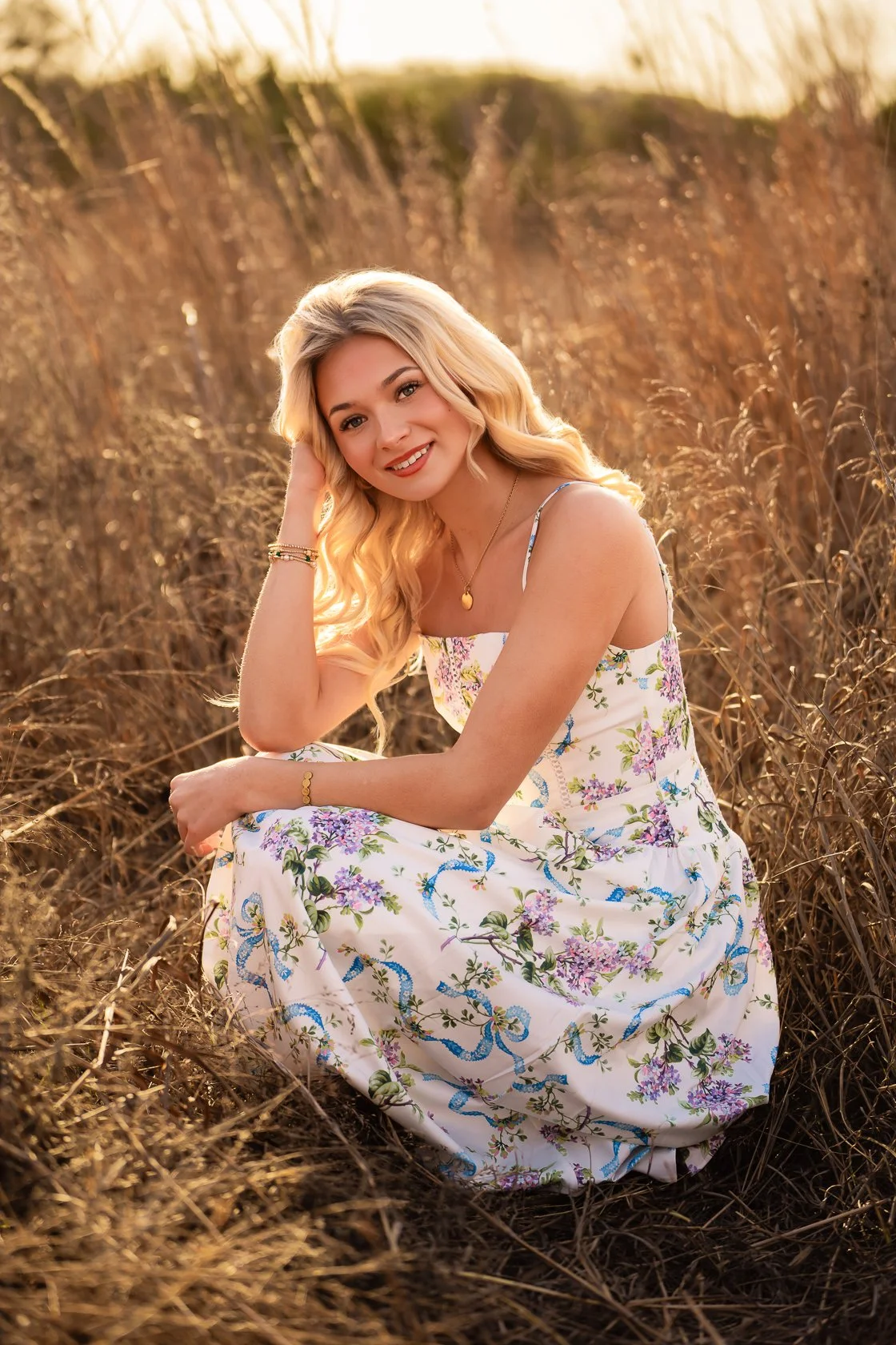 A woman with long blonde hair, wearing a white floral dress, sitting in a field of tall dry grass during sunset, smiling at the camera.
