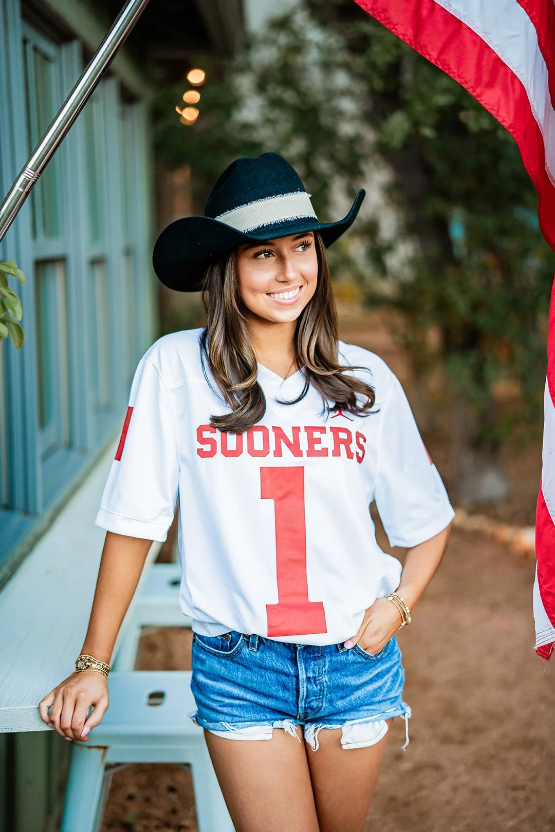 Young woman in a University of Oklahoma football jersey, denim shorts, and a cowboy hat, smiling outdoors near an American flag.