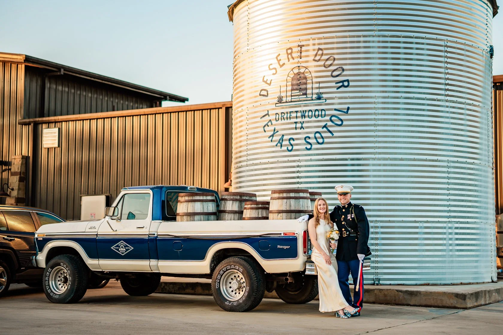 A couple in formal wedding attire standing beside a vintage blue and white pickup truck with barrels in the truck bed, next to a large metal structure with a sign that reads "Desert Door TexassS" in Driftwood, Texas.