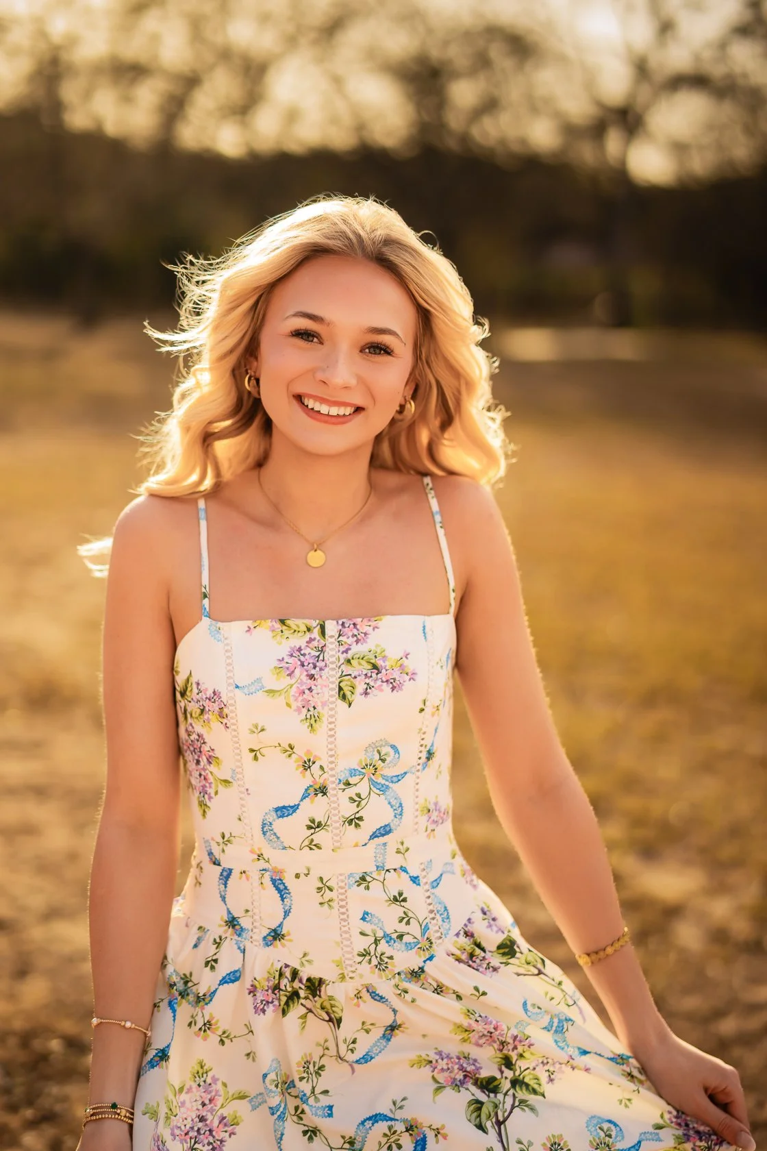 A young woman with blonde curly hair smiling outdoors in a floral summer dress during golden hour