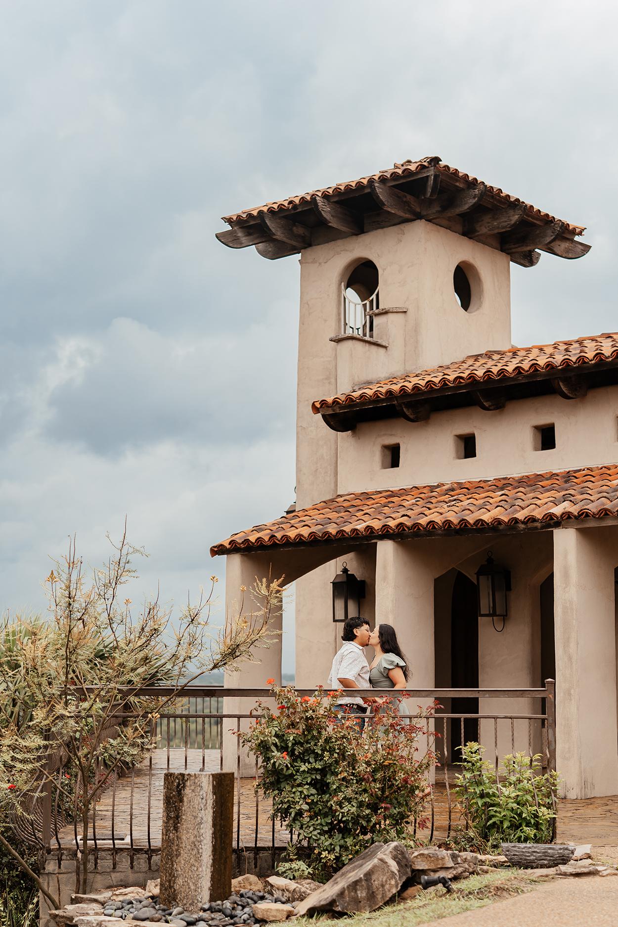 A couple sharing a kiss on a balcony of a Spanish-style building with tiled roof, stone walls, and a small bell tower, under a cloudy sky.