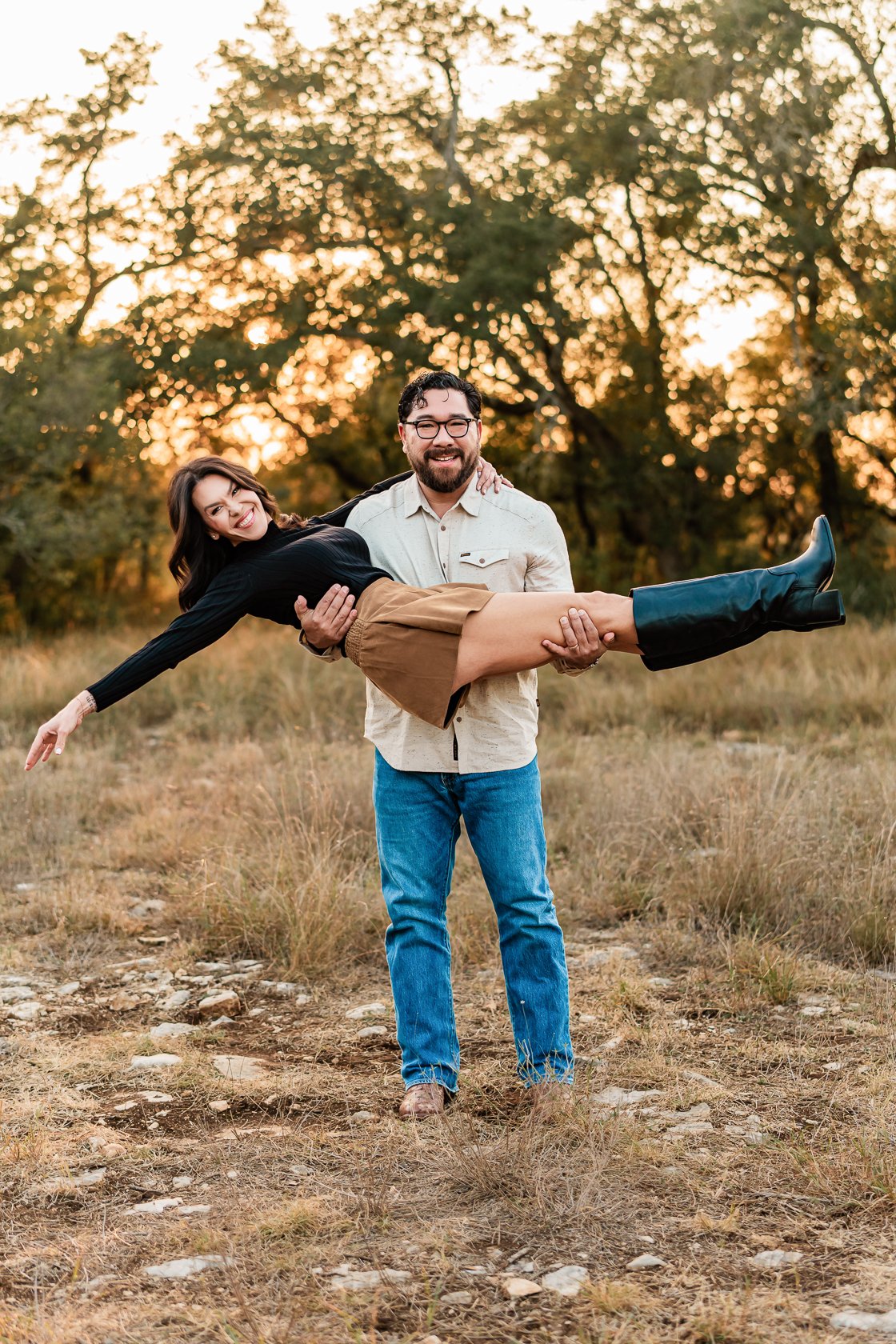 A man with glasses and a beard holding a woman in a black top, tan skirt, and black boots in a field during sunset.
