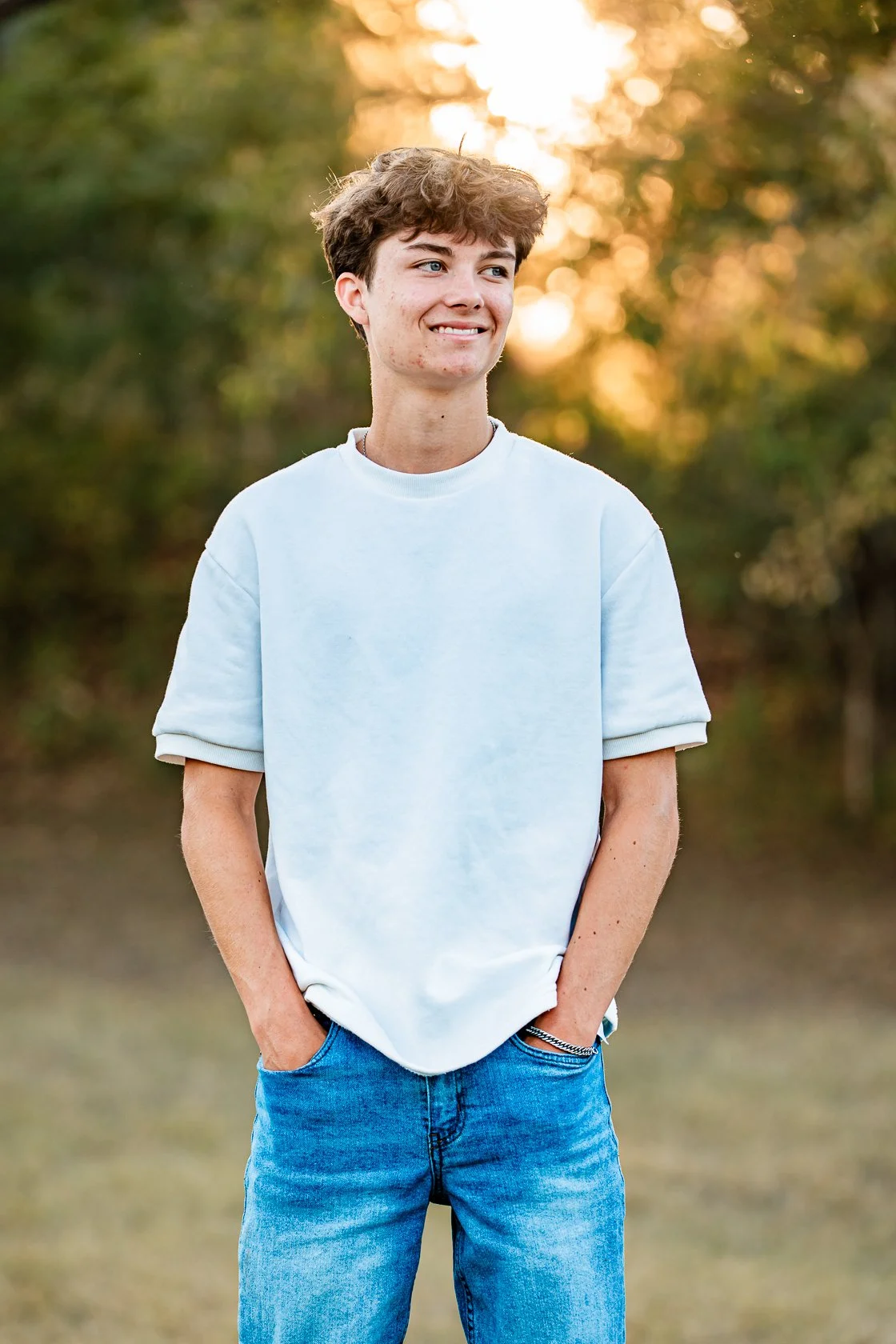 A young man with brown, curly hair stands outdoors with hands in his pockets, smiling and looking to the side, with trees and the setting sun in the background.