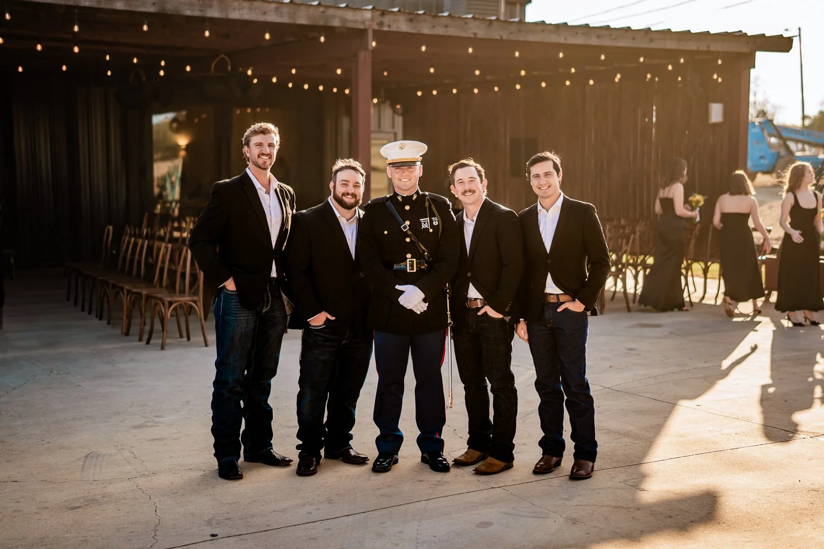 Group of five men standing outside in the late afternoon, with a wooden building with string lights behind them. One man is in a decorated military uniform and the other four are in black suits with white shirts.