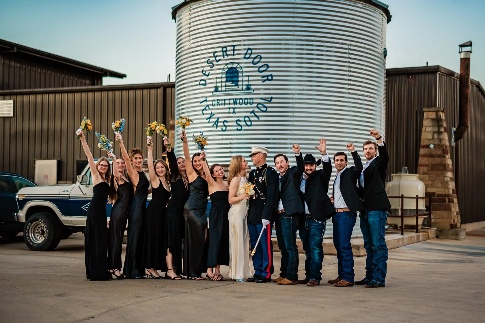Group of people celebrating a wedding outdoors near a large silo with a sign that reads 'Desert Door Tacos Thas So.' The bride and groom are in the center, with the bride holding a bouquet and the groom in a military uniform. The group includes women