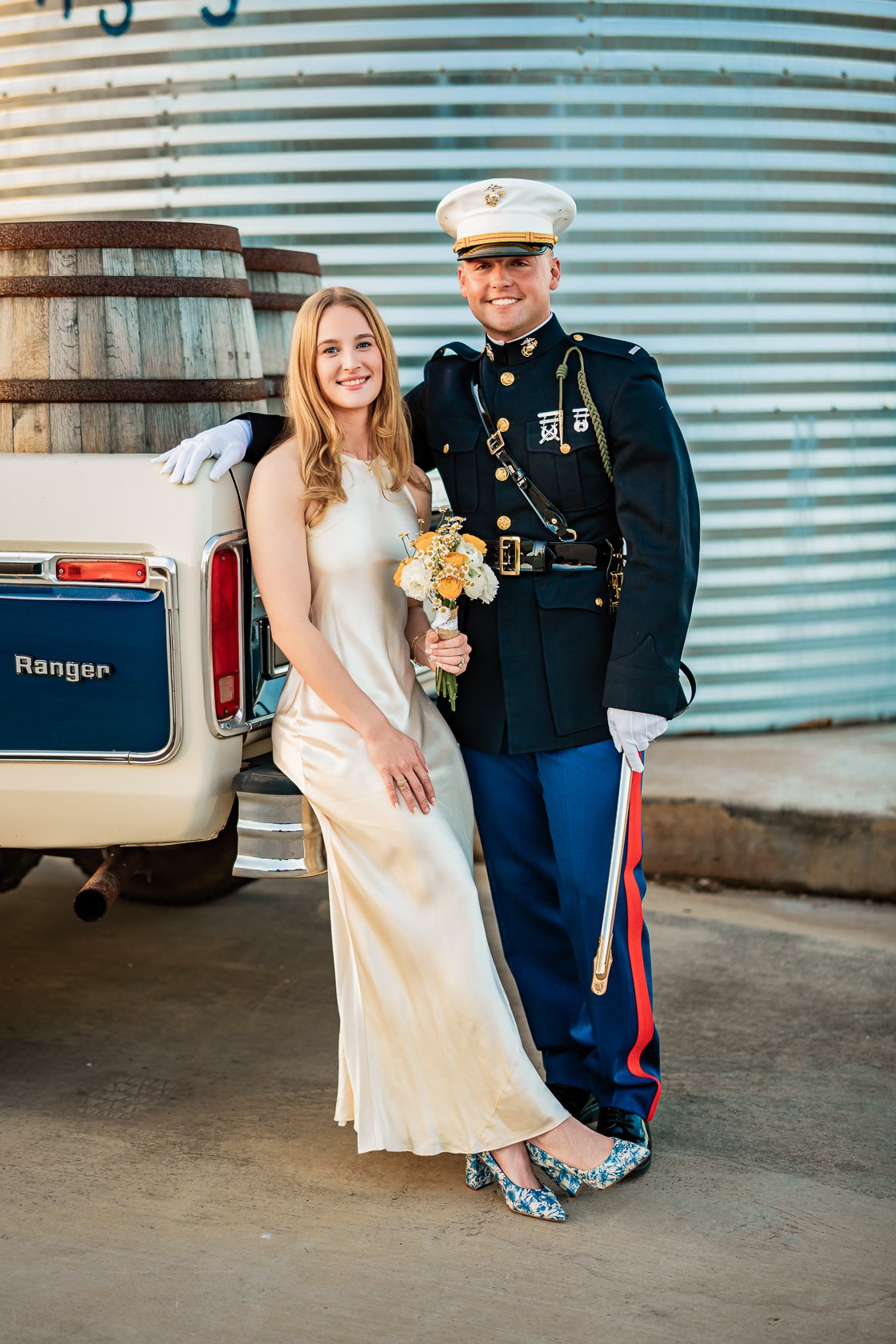 A smiling couple poses outdoors in front of a vintage white car and a metal building. The woman, holding a small bouquet, wears a cream-colored satin dress and floral heels. The man, in a U.S. military dress uniform, stands beside her with his arm ar