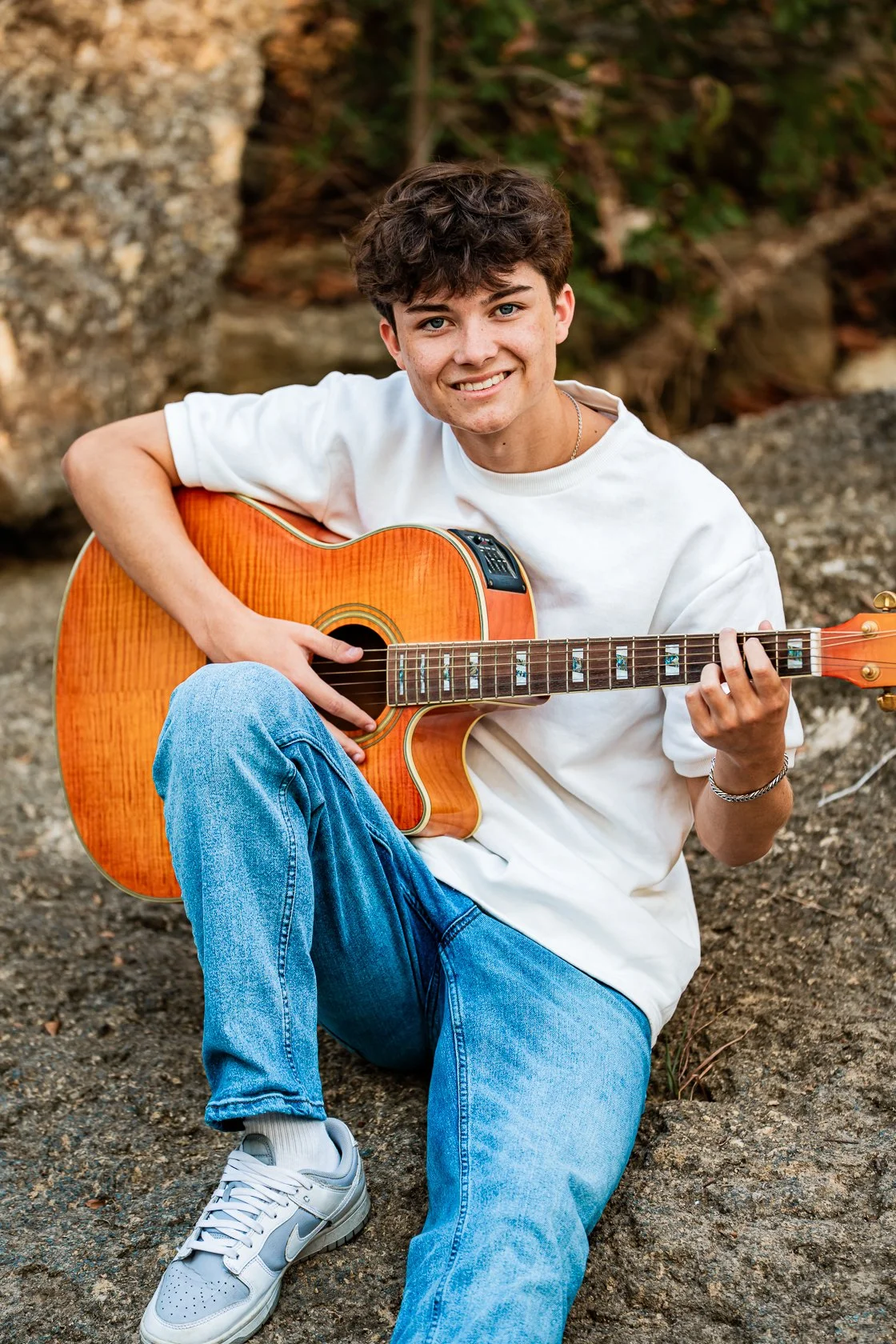 A young man with curly brown hair, wearing a white T-shirt, blue jeans, and white Nike sneakers, sitting on the ground outdoors while playing an orange acoustic guitar and smiling at the camera.