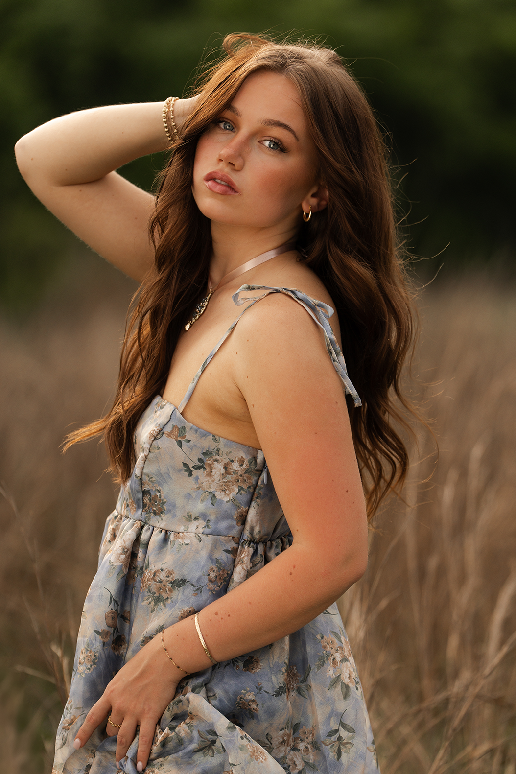 A young woman with long brown hair, wearing a floral dress, standing in a field with tall grass, with a green, blurred background, looking at the camera.