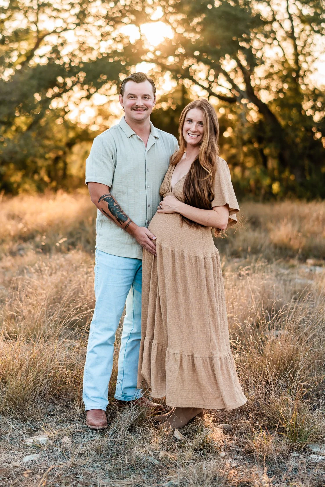 A couple standing outdoors in a field during sunset, with the woman holding her pregnant belly and the man touching her belly, both smiling.