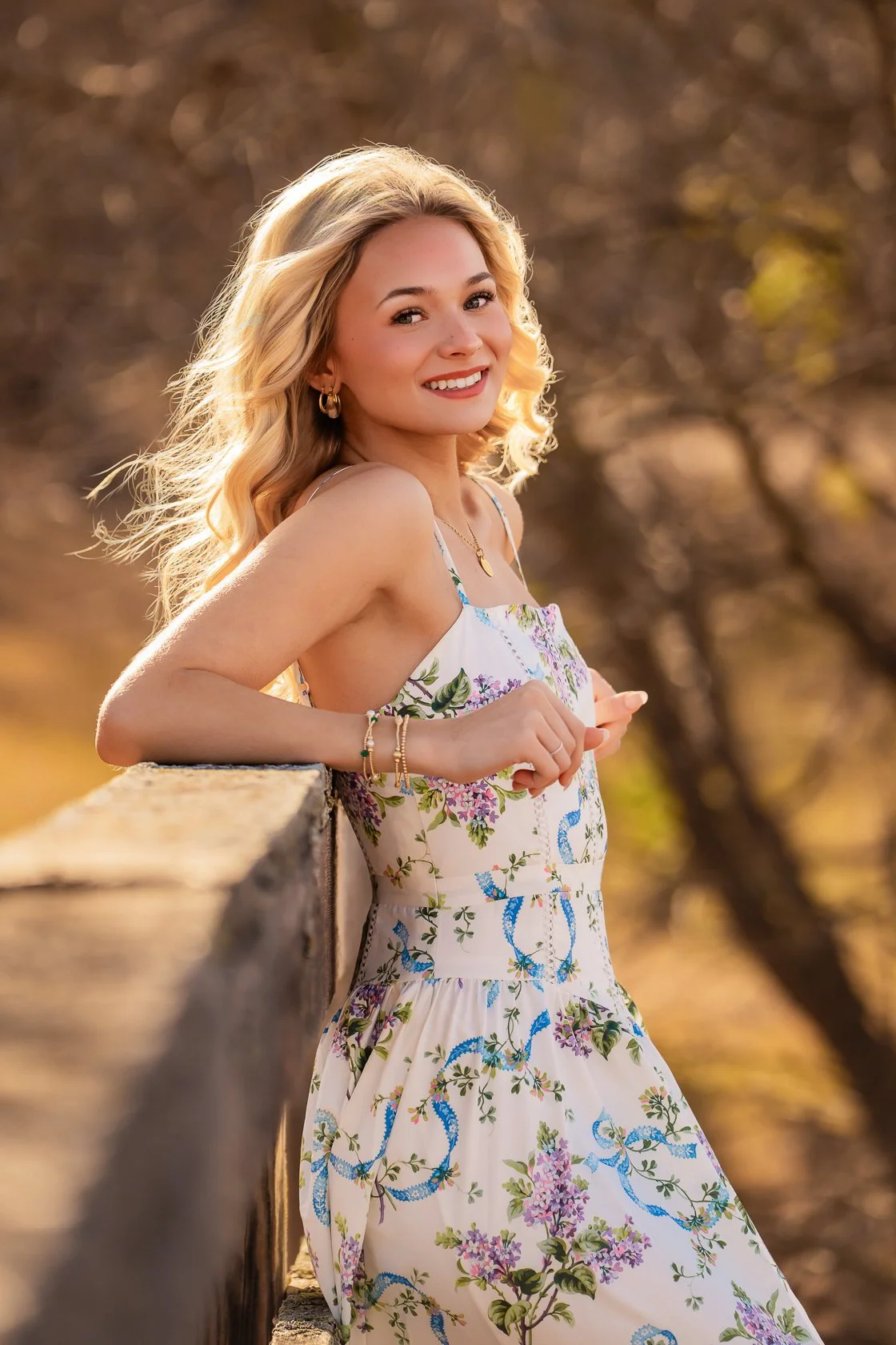 A young woman with long, wavy blonde hair smiling, leaning on a wooden fence outdoors during sunset, wearing a white floral sundress and jewelry.