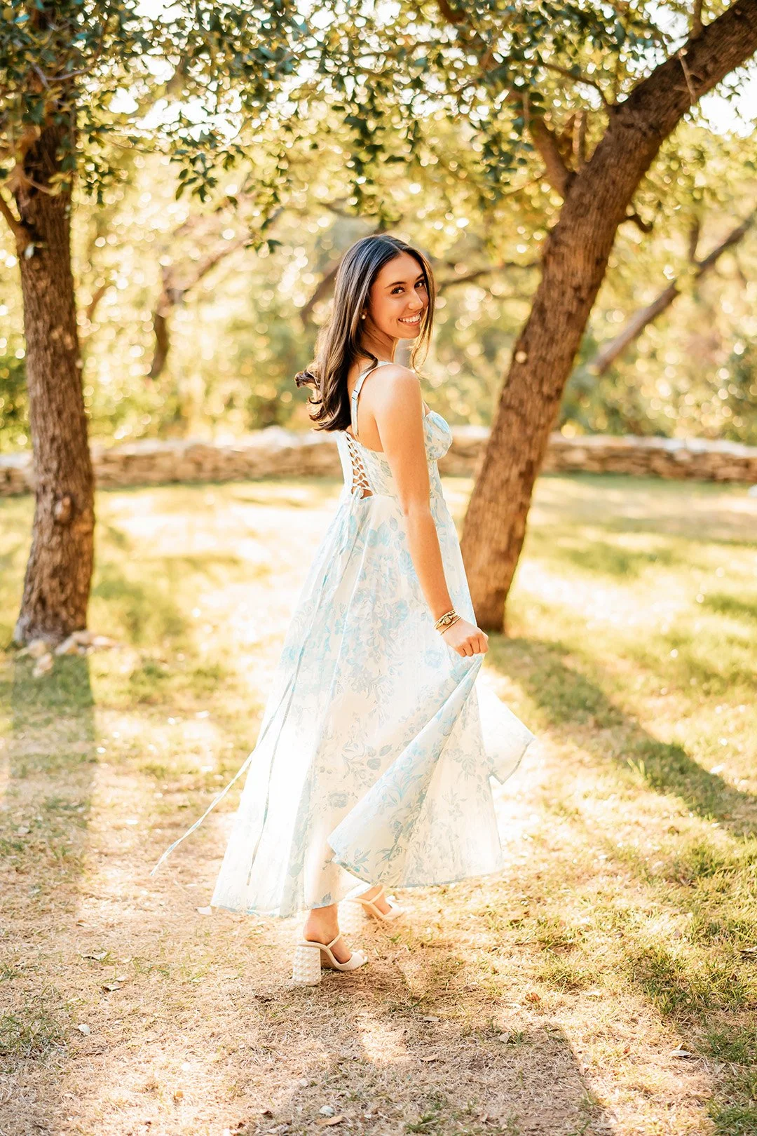 A woman in a light-colored floral dress outdoors under trees, smiling at the camera, holding up her dress slightly while standing on a grassy path.