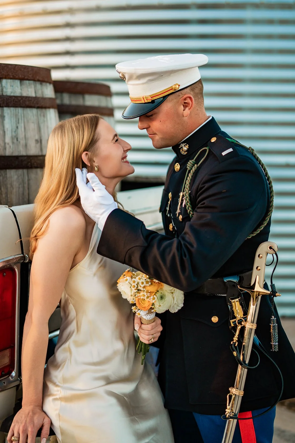 A smiling woman in a white dress holding a bouquet of flowers is being tenderly touched on the face by a man in a military uniform. They are standing outdoors near a car, with a wooden barrel and a corrugated metal wall in the background, sharing an 