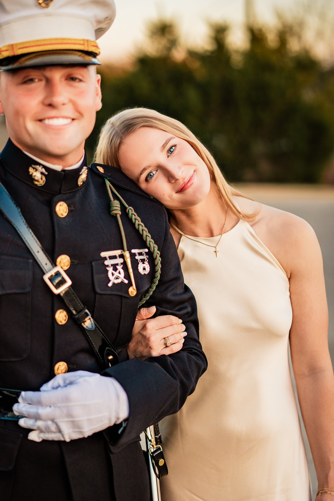 A woman with blonde hair leaning her head on a man in a military uniform, both smiling softly outdoors at sunset.