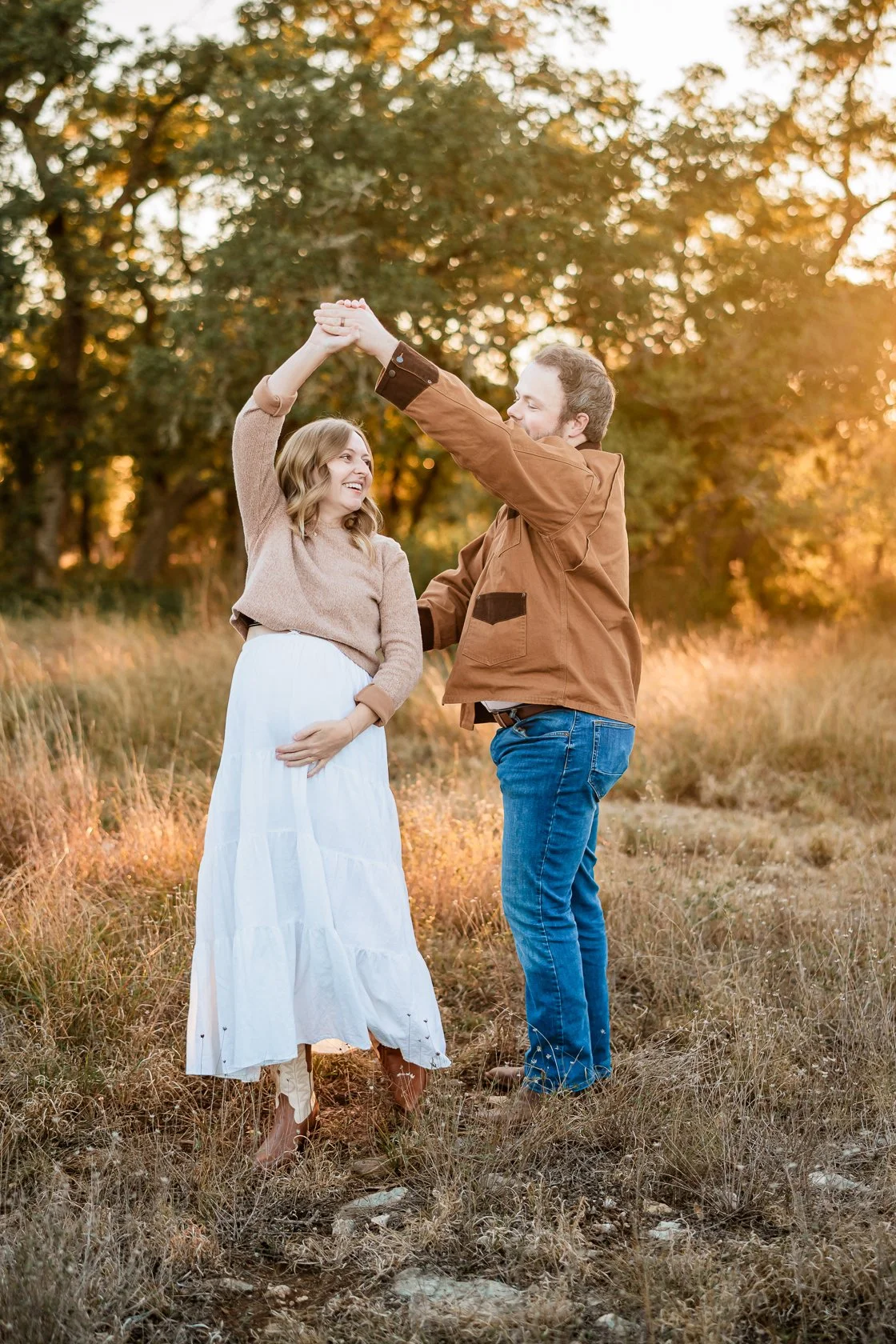 A man and woman dancing outdoors during sunset, with trees in the background, both smiling, holding hands up, woman in a white skirt and brown sweater, man in a brown jacket and blue jeans.
