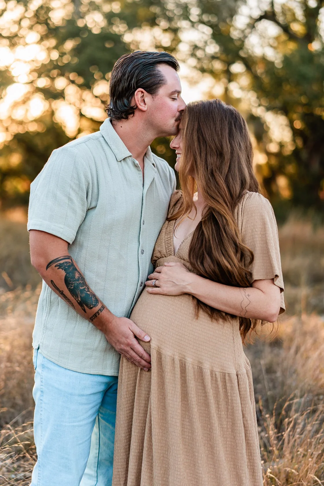 A man kisses a pregnant woman on her forehead in a field during sunset, with trees and warm lighting in the background.