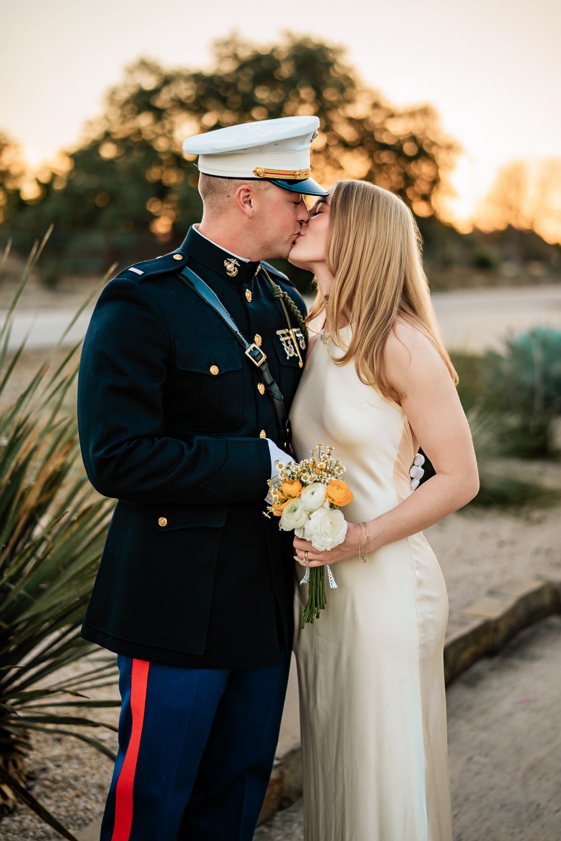 A couple kissing outdoors at sunset, the man dressed in a formal military uniform and the woman in a white dress holding a bouquet of flowers.