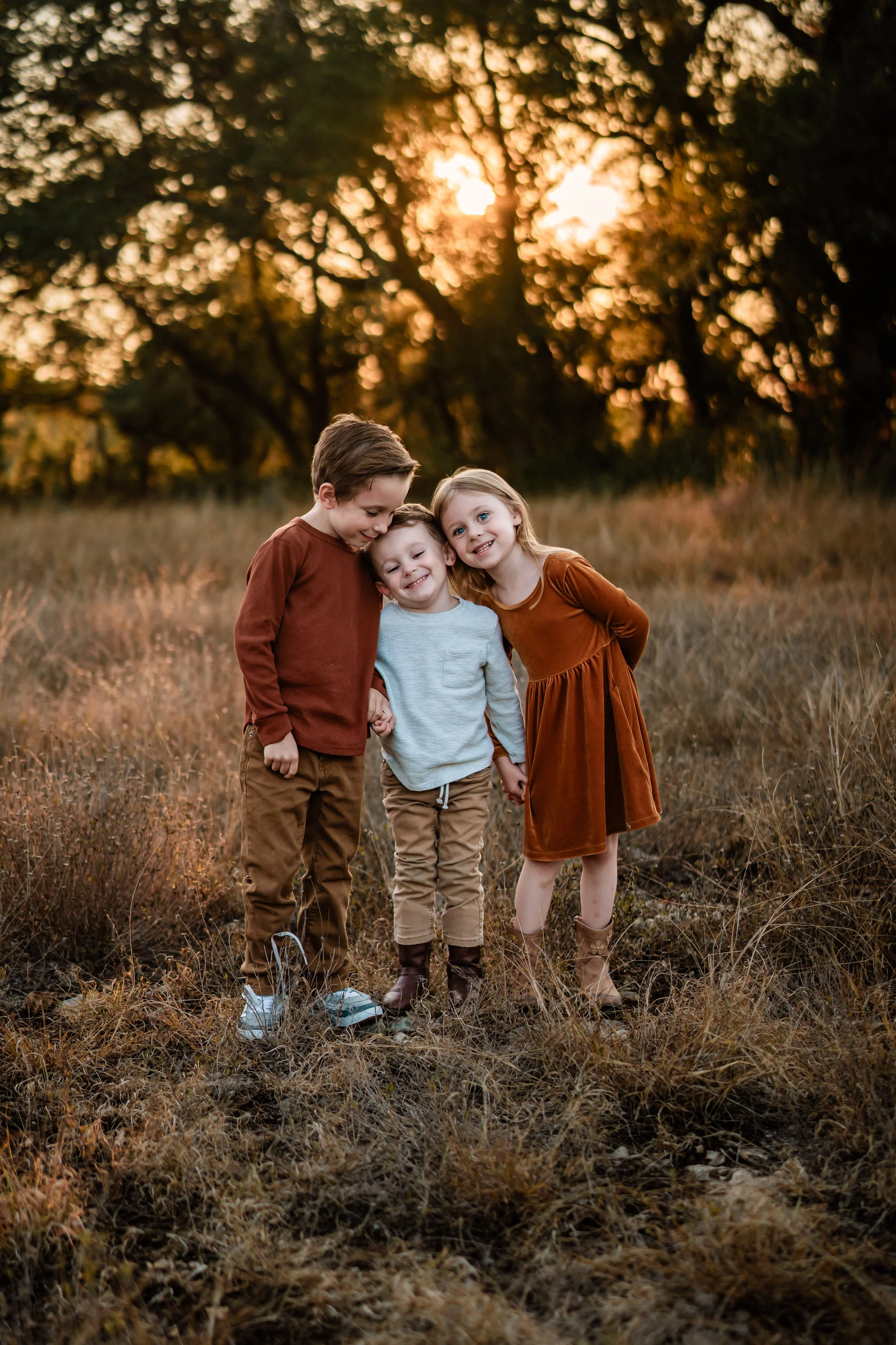 Three children holding hands in a field at sunset, smiling and leaning on each other.