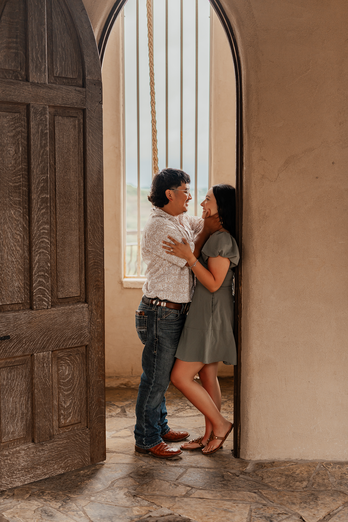 A couple standing in a doorway, smiling and touching faces, with a window and rope behind them.