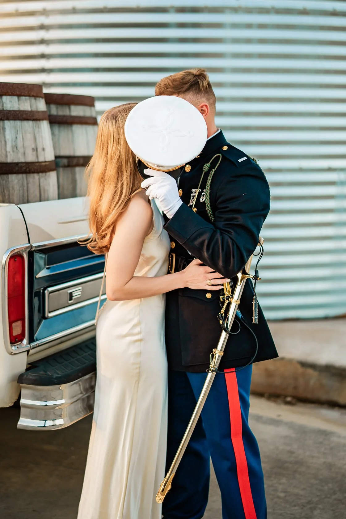 A bride and a military officer share a kiss, with the bride in a cream-colored dress and the officer in a formal military uniform, standing outdoors near a vintage white vehicle and metal siding.