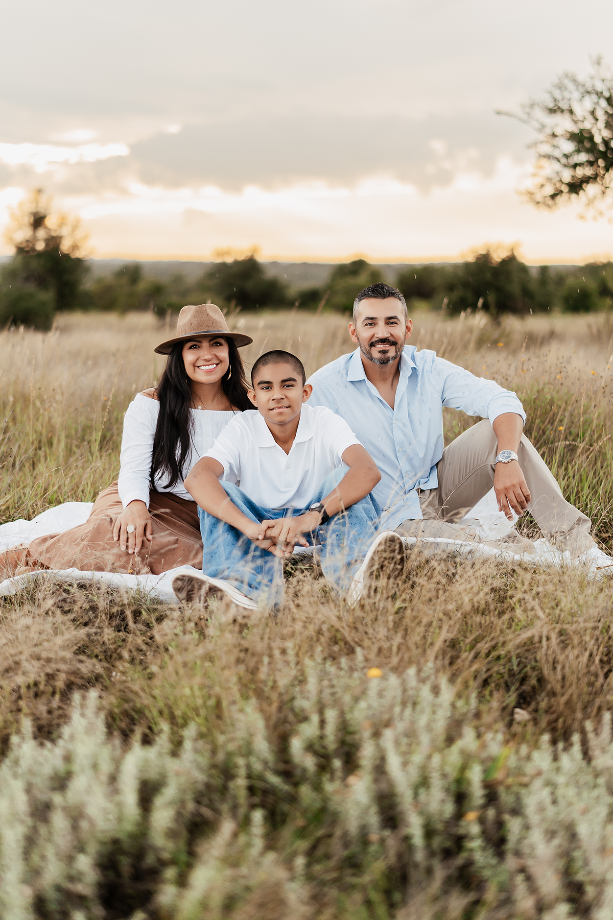 A family of three sitting on a blanket in a field during sunset, smiling at the camera.
