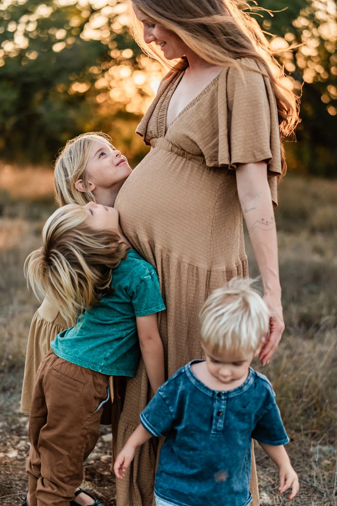 A pregnant woman in a tan dress is surrounded by four children in an outdoor setting during sunset. The children are hugging and looking lovingly at her.
