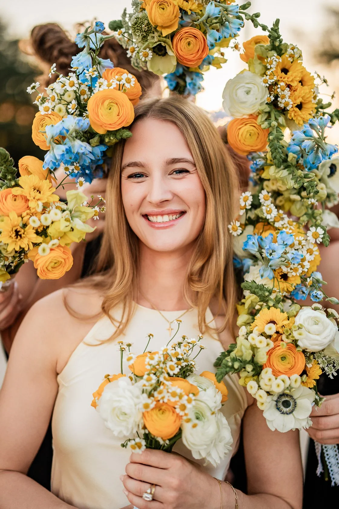 A smiling woman with wavy, shoulder-length blond hair holding a bouquet of white, yellow, and orange flowers in front of her chest. She is surrounded by other women holding floral arrangements and wearing floral crowns, with trees and a sunset in the