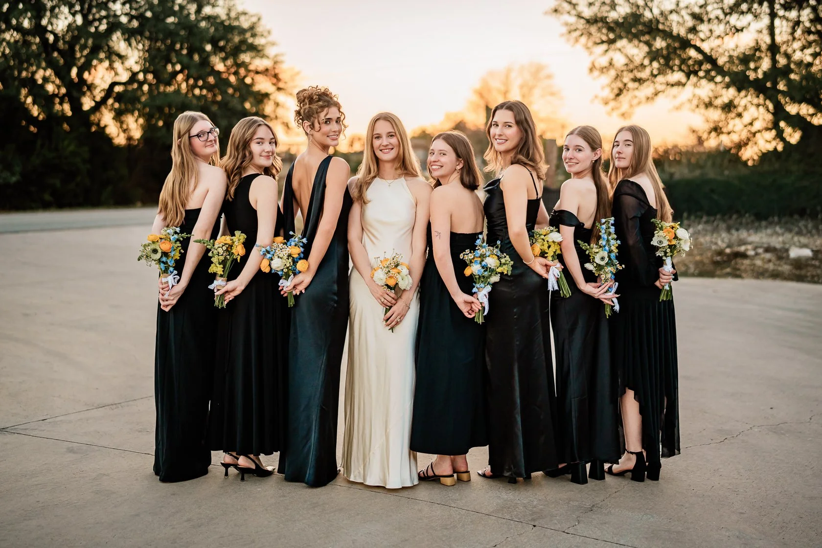 A group of nine women, including a bride in a white dress, posing outdoors during sunset. The women are dressed in black, holding floral bouquets, and are standing on a wide open space with trees in the background.