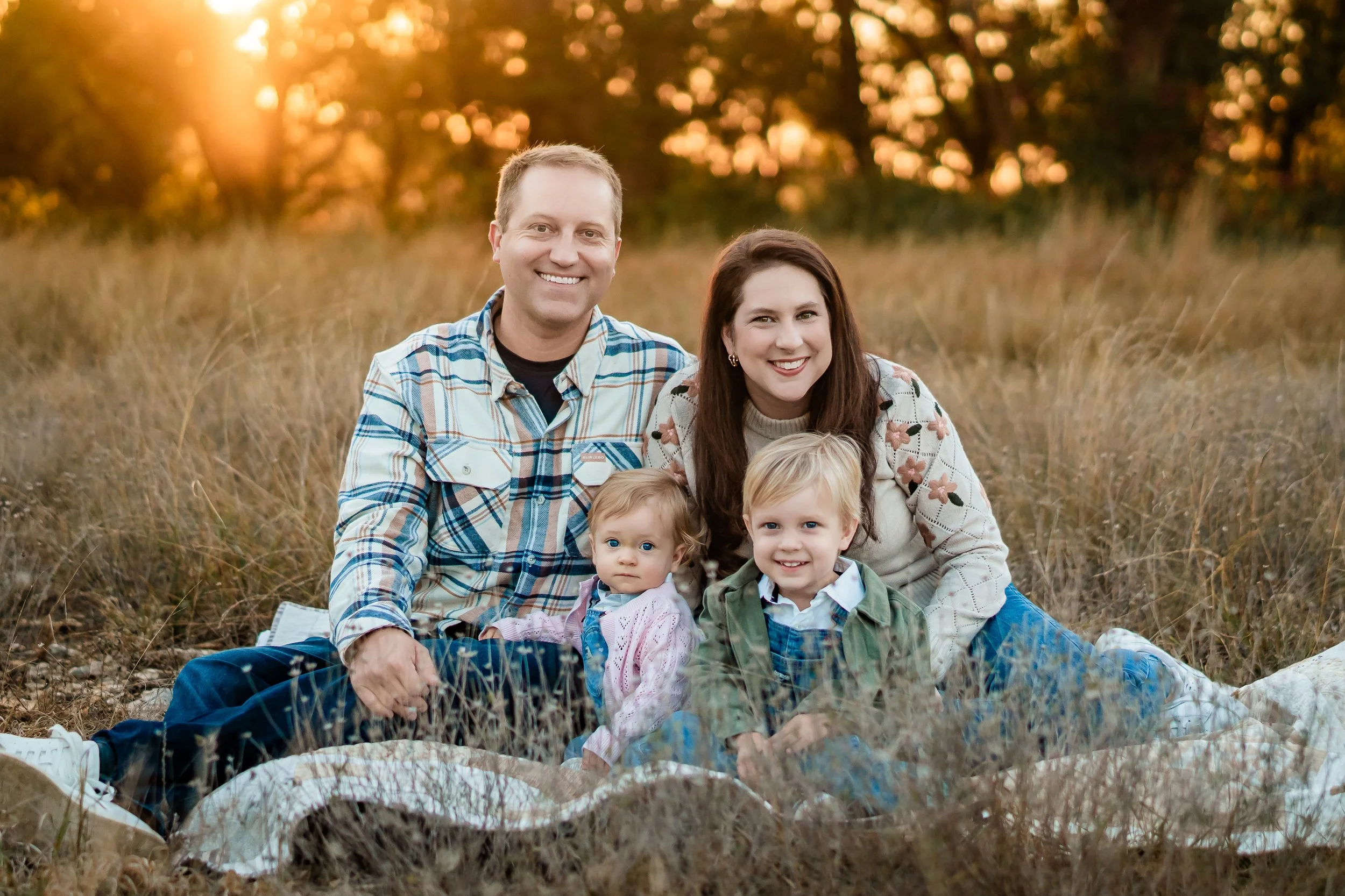 A happy family of four sitting on a blanket in a field during sunset, smiling at the camera.