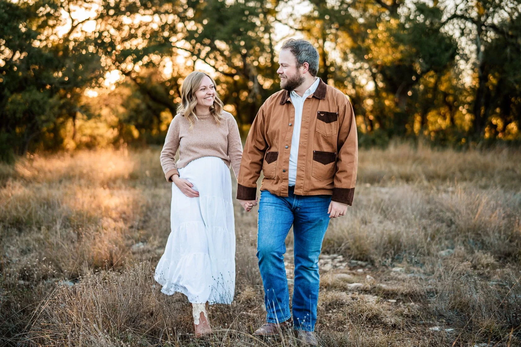 A couple walking hand in hand through a field at sunset. The woman is pregnant and wearing a beige sweater and white skirt, while the man is dressed in a brown jacket and blue jeans. They are smiling at each other with trees in the background.